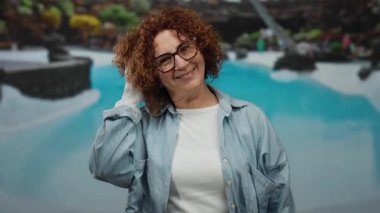 Gülümseyen orta yaşlı bir kadın, gözlüklü, açık hava otelinin havuzunda kıvırcık saçlarına dokunuyor..
