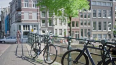 Amsterdam 'daki bir kanal köprüsündeki bisiklet manzarası bulanık. Dağılmış yayalar ve renkli tarihi binalar Hollanda' daki kentsel yaşamın özünü yansıtıyor..