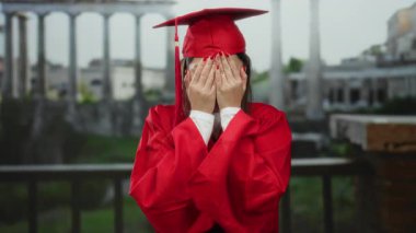 Kırmızı mezuniyet elbisesi giymiş bir kadın antik Roma harabelerinin önünde sevinçle poz veriyor. Arka planda bulanık bir şekilde kutlama yapıyor ve tarihi bir manzara sergiliyor.