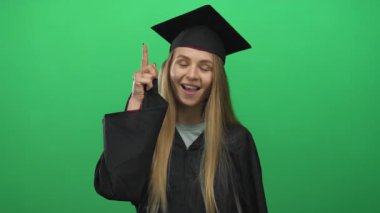 Mezuniyet üniformalı genç beyaz kadın yeşil arka planda kutlama yapıyor, neşeli bir ifadeyle, başarıyı ve başarıyı sembolize ediyor..