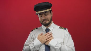 Pilot üniforması giyen genç sakallı İspanyol adam ellerini göğsüne koyuyor. Kırmızı bir arka plana doğru. Duygularını ve profesyonelliğini ifade ediyor..