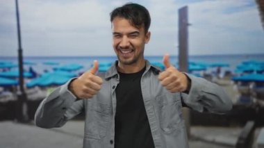 Mavi şemsiyeli ve arka planda sakin okyanus sularıyla pitoresk bir sahilde baş parmağıyla gülümseyen İspanyol genç adam..