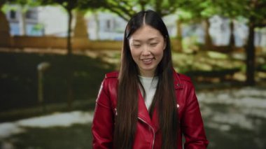 Parkta gülümseyen kırmızı ceketli bir kadın, güneşli bir günde, bulanık yeşil bir arka planda uzun saçlarını gösteriyor..