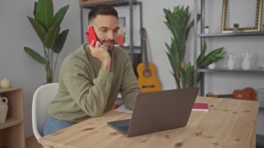 Kırmızı telefonda konuşan sakallı genç adam yeşil bitkilerle dolu oturma odasında dizüstü bilgisayar kullanıyor gitar ve ahşap raflar konsantrasyon ve iletişim gösteriyor..