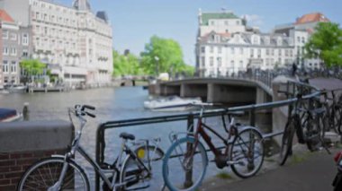 Bisikletler, bulanık şehir manzarası ve uzaktaki tarihi binalarla dolu kanal parmaklıkları boyunca dizilmiş, tipik bir Hollanda kentsel manzarasını gözler önüne seriyor.