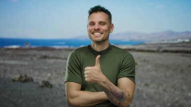 Dövmeleri olan genç bir adam, güneşli bir sahilde güvenle gülümsüyor, okyanusla rahat bir atmosferi kucaklıyor ve arka planda berrak bir gökyüzü, sakin bir sahil atmosferi sergiliyor..