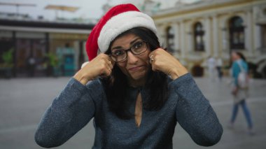 Gözlüklü kadın kırmızı Noel Baba şapkası takıyor ve şehir binasının önünde yumruklarını yanaklarına bastırıyor; bayram nostaljisi üzüntüsü pişmanlığı.