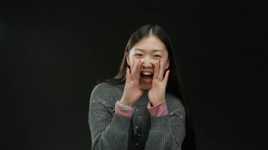 Siyah arka planda sıcak süveter giyen, uzun saçlı, neşeli yüzlü, neşeli bir kadın tezahürat ve coşkuyla bağırıyor..