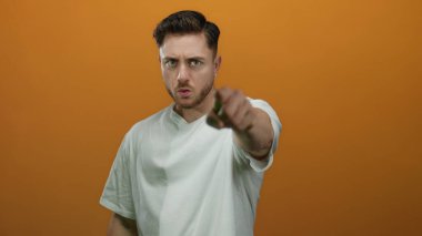 Sakallı genç adam, izole edilmiş turuncu arka planda kameraya kızgın bir şekilde el kol hareketi yapıyor. Günlük beyaz gömlek giyiyor..