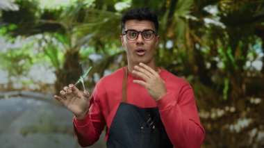 Parkta gözlüklü ve önlüklü genç İspanyol bir adam, kendinden emin bir şekilde bir kuaförün karizmasını betimliyor açık hava yeşilliğinin ortasında, modern stil ve beceri yayıyor..