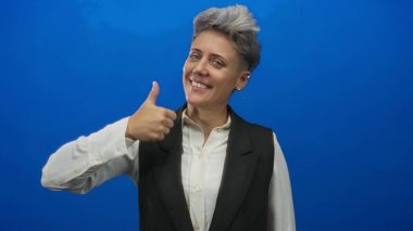 Mavi stüdyoda beyaz gömlek giyen, siyah yelek giyen, gülümseyen ve baş parmak işareti yapan bir kadın..