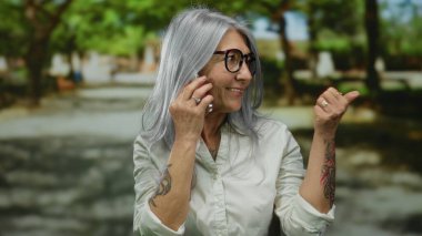 Sunny Park 'ta gülümseyen ve el kol hareketi yapan, günlük kıyafetler giyen ve dövme gösteren yaşlı gri saçlı kadın, canlı ve çekici bir açık hava sahnesi yaratıyor..