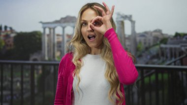 Açık havada pembe daireler çizen bir kadın. Yüzüne yakın el hareketleriyle antik harabelerin önünde duruyor. Sütunları ve doğası olan canlı bir açık hava ortamında şaşkınlık ve neşe ifade ediyor..