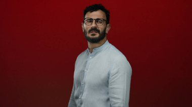 Sakallı ve gözlüklü İspanyol bir adam sıradan bir gömlekle koyu kırmızı bir arka plana karşı duruyor. Kendine güveni ve tarzı var..