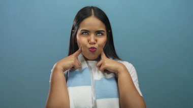 Mavi eşarplı genç bir kadın soyutlanmış mavi arka planda komik suratlar yapıyor, neşeli ifadeler ve duygular sergiliyor..