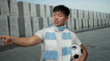 Beyaz tişörtlü ve atkılı genç Çinli adam şehir caddesinde futbol topu tutarken enerjik bir şekilde dışarıyı gösteriyor..