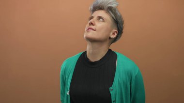 Yeşil hırkalı ve siyah küpeli bir kadın turuncu stüdyo ortamında başını eğiyor..