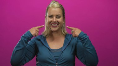 Kadın, pembe stüdyoda mavi eşofman üstü giyerek geniş bir gülümseme sergilerken parmaklarını dişlerine işaret ediyor..