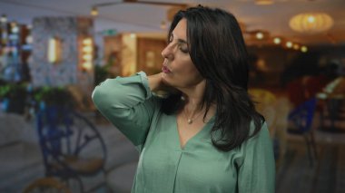 Orta yaşlı, boynunda jest olan İspanyol bir kadın ve otel lobisinde gözleri kapalı, hafifçe aydınlatılmış oturma alanı var..