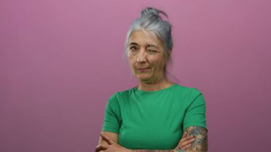 Gri saçlı, dövmeli, kolları çapraz parlak pembe arka planı olan son sınıf öğrencisi bir kadın..