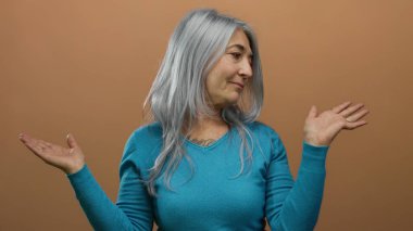 Mavi kazaklı yaşlı kadın, izole edilmiş kahverengi arka plan duvarına el hareketi yaparak rahat ve düşünceli bir davranış sergiliyor..