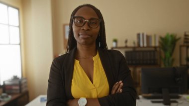 Sarı gömlekli ve gözlüklü bir kadın, arka planda bilgisayar, raf ve belgelerle profesyonel çalışma alanını sergiliyor..