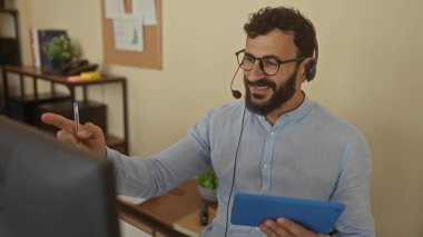 Ofis ortamında, raflar ve arkaplanda modern çalışma alanlarını yansıtan grafiklerle çevrimiçi toplantı yaparken, elinde tablet tutan sakallı İspanyol bir adam..