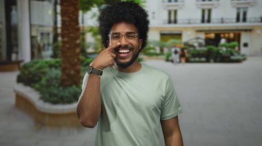 Gülümseyen adam, yeşil bitkiler ve arka planda binalar olan, gözlüklü ve günlük kıyafetlerle şehir dışında duran burnu işaret ediyor..