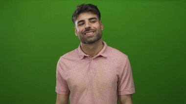Pembe polo tişörtlü genç İspanyol adam yeşil stüdyo ortamında hafifçe başını eğerek gülümsüyor..