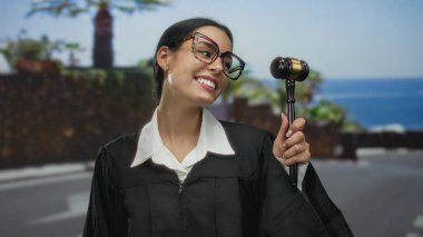 Gülümseyen kadın yargıç elinde tokmağıyla dışarıda tropikal bir zemin tutarak coşkulu profesyonelliğini ve kültürel mirasını canlı bir yasal ortamda sergiliyor..