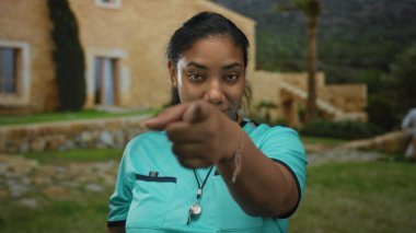 Turkuaz formalı hakem kadın ıslık çalıyor ve güneş ışığı altında taş cepheli bir binanın girişinde kamerayı işaret ediyor..