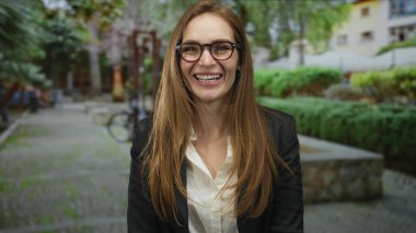 Gözlüklü ve siyah ceketli kadın, yeşil parktaki taş bankta otururken kameraya geniş bir şekilde gülümsüyor. Mutluluk..