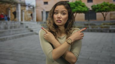 Uzun saçlı, açık havada sırıtan bir kadın iki parmağıyla işaret ederken güneşli bir avluda günlük kıyafetler ve kol saati takıyor..
