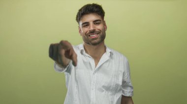 Gülümseyen genç İspanyol adam açık yeşil stüdyo alanında beyaz gömlek giyerken doğrudan kamerayı işaret ediyor..