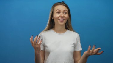 İzole edilmiş mavi arka plana karşı beyaz gömlekle gülümseyen genç ve anlamlı duygular taşıyan kadın jestleri.