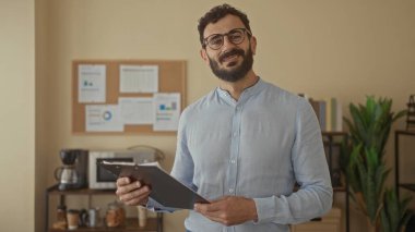 Sakallı, pano tutan İspanyol bir adam modern ofiste arka planda çizelgeleri olan rahat tavırlı profesyonel iç mekan düzeniyle duruyor..