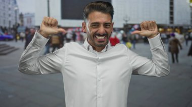 Hispanik adam geniş bir sırıtıyor ve şehir merkezinde hareketli bir caddede başparmaklarını göğsüne işaret ediyor; kendine güven.