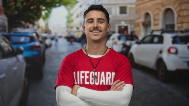 Kırmızı tişörtlü şehir caddesinde kolları çaprazlanmış genç İspanyol cankurtaran güneşli bir günde arka planda bulanık şehir arabalarıyla güvenle gülümsüyor..