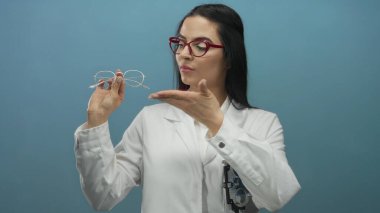Gözlüklü ve üniformalı kadın gözlükçü, profesyonel hizmeti vurgulayan mavi arka plan duvarına karşı gözlük tutuyor.