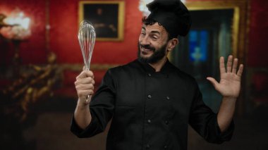 Şef gibi giyinmiş İspanyol sakallı bir adam sanat galerisinde çırpıcıyla kırmızı duvarlar ve dekoratif çerçevelerle müze ortamında aşçılık hevesi sergiliyor..