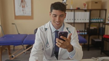 Steteskoplu beyaz önlüklü doktor akıllı telefon tutuyor ve klinik masasında ekranı dinliyor. Merhamet görevi..