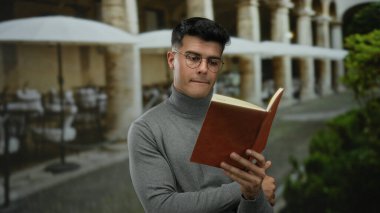 Gözlüklü genç adam, yeşil ve şemsiyelerle çevrili bir açık şehir restoranının terasında sakin ve çalışkan bir ortam sağlayan bir kitap okuyor..