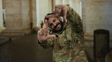 Askeri üniformalı İspanyol bir adam, eski Roma şehir ortamında gülümseyen elleriyle manzara çerçevesi çiziyor, hikaye anlatma ya da fotoğrafçılık öneriyor..