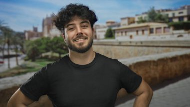 Güneşli bir günde, güzel bir mimari ve yemyeşil ile çevrili, canlı bir atmosferi olan tarihi bir Akdeniz şehrinde, açık havada gülümseyen genç sakallı adam..