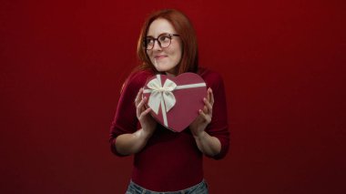 Kadın gülümserken kalp şeklinde hediye kutusunu kırmızı arka planda tutarken aşk ve romantizm kavramını zarafet ve stil ile gösteriyor.