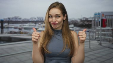 Mavi bluzlu kadın deniz kenarı iskele inşaatında iki eliyle baş parmağını kaldırıyor. Onaylama iyimserliği.