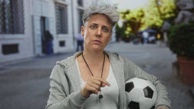 Kol altında futbol topu tutan gri kapüşonlu kadın koç parmaklarını sokakta öne uzattı; otorite disiplin motivasyon liderliği.