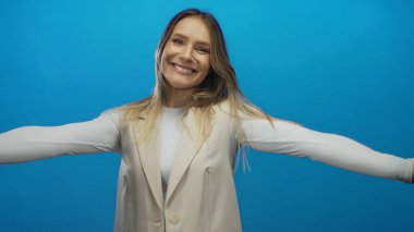 Mavi izole edilmiş arka plana karşı kollarını açmış gülümseyen kadın mutluluk ve sıcaklık yayıyor, pozitiflik, neşe ve dostça selamlaşma kavramları için ideal..