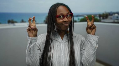 Kıvırcık saçlı bir kadın deniz kenarında gezinirken tırnak işareti yapıyor. Plaj zemininde gözlük takıyor ve bulutlu bir gökyüzünün altında rahat bir ifade takınıyor..
