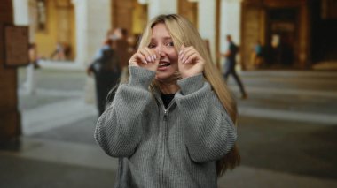 Gri kazaklı sarışın genç bir kadın açık hava üniversitesi ortamında korkmuş görünüyor. Arka planda eski bir mimari, kamusal alanı gösteriyor..